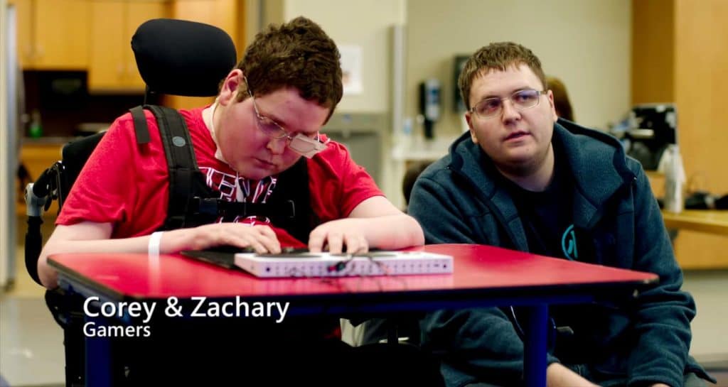 Dois meninos brancos, de cabelos claros, estão sentados ao redor de uma mesa de tampo vermelho. O menino da esquerda usa um óculos com tampão no olho esquerdo, está numa cadeira de rodas e manuseia o Adaptive Controller disposto na mesa à sua frente. O menino da direita está olhando para frente.
