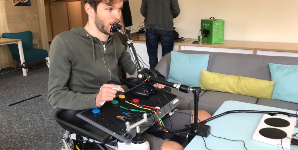 Foto de um homem branco de barba sentado na cadeira de rodas. A sua frente um pedestal com um controle operado pela boca. Em seu colo há uma bandeja com botões grandes e coloridos ao lado de um controle de Xbox, ligados por um fio a um Adaptive Controller.