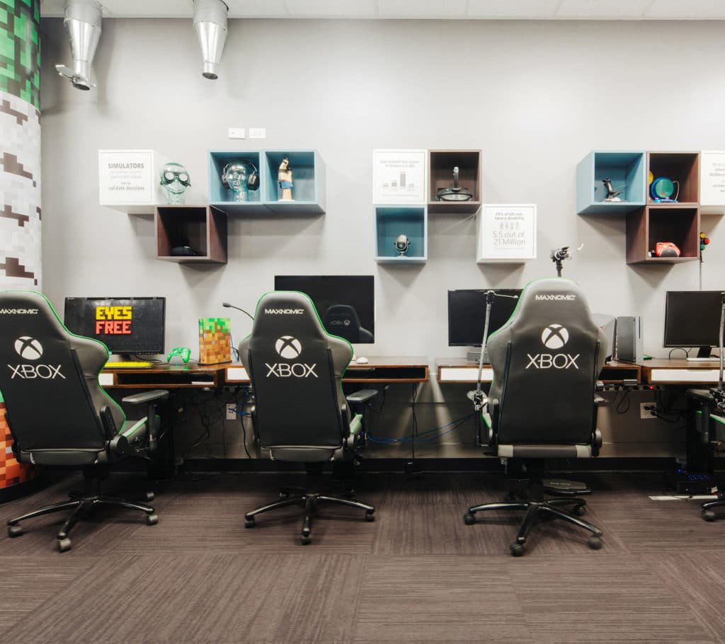 Sala de paredes cinzas e chão de carpete. Na parede há 4 estações formadas por mesa com monitor e uma cadeira com o logo do xBox no verso. O primeiro monitor a esquerda está com a tela coberta por uma proteção que diz Eyes Free.