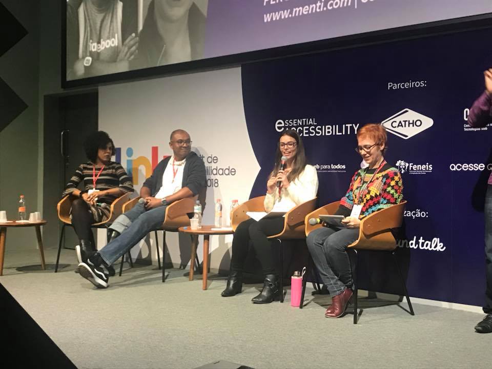 As quatro cadeiras do palco do evento estão ocupadas pelos palestrantes do painel, da esquerda para a direita: Claudiene Mortimer, uma mulher negra de cabelos cacheados e vestido preto listrado, Alexandre Theodoro, um homem negro, de óculos, camiseta branca com um casaco cinza, calça jeans e uma prótese em uma das pernas, Milena Delfine, uma mulher branca de cabelos lisos castanhos, veste uma blusa branca e uma calça preta e Ivone Santana, uma mulher branca de cabelos ruivos curtos, usa uma bata colorida e calça jeans e segura o microfone.