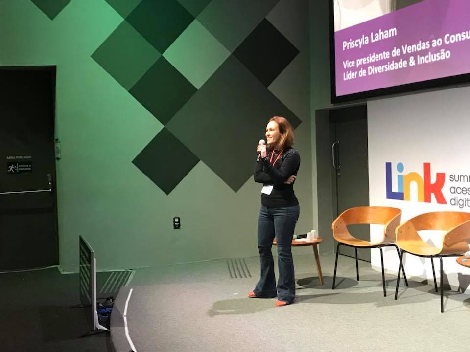 Priscyla Laham, uma mulher branca de cabelos loiros e roupa preta, está em pé no palco e segura o microfone na altura da boca.