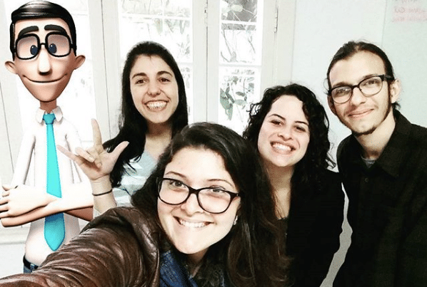 A foto mostra quatro pessoas tirando uma selfie e, à esquerda delas, o Hugo, personagem virutal, de braços cruzados (inserido digitalmente na foto). Da esquerda para direita, uma menina de cabelos pretos e pele branca sorrindo e fazendo o sinal de amor em Libras, uma menina de cabelos castanhos, pele branca e óculos sorrindo, a Ana Clara, de pele branca, calos pretos e roupa preta sorrindo e um homem de cabelos grandes, óculos, barba rala e roupa preta.