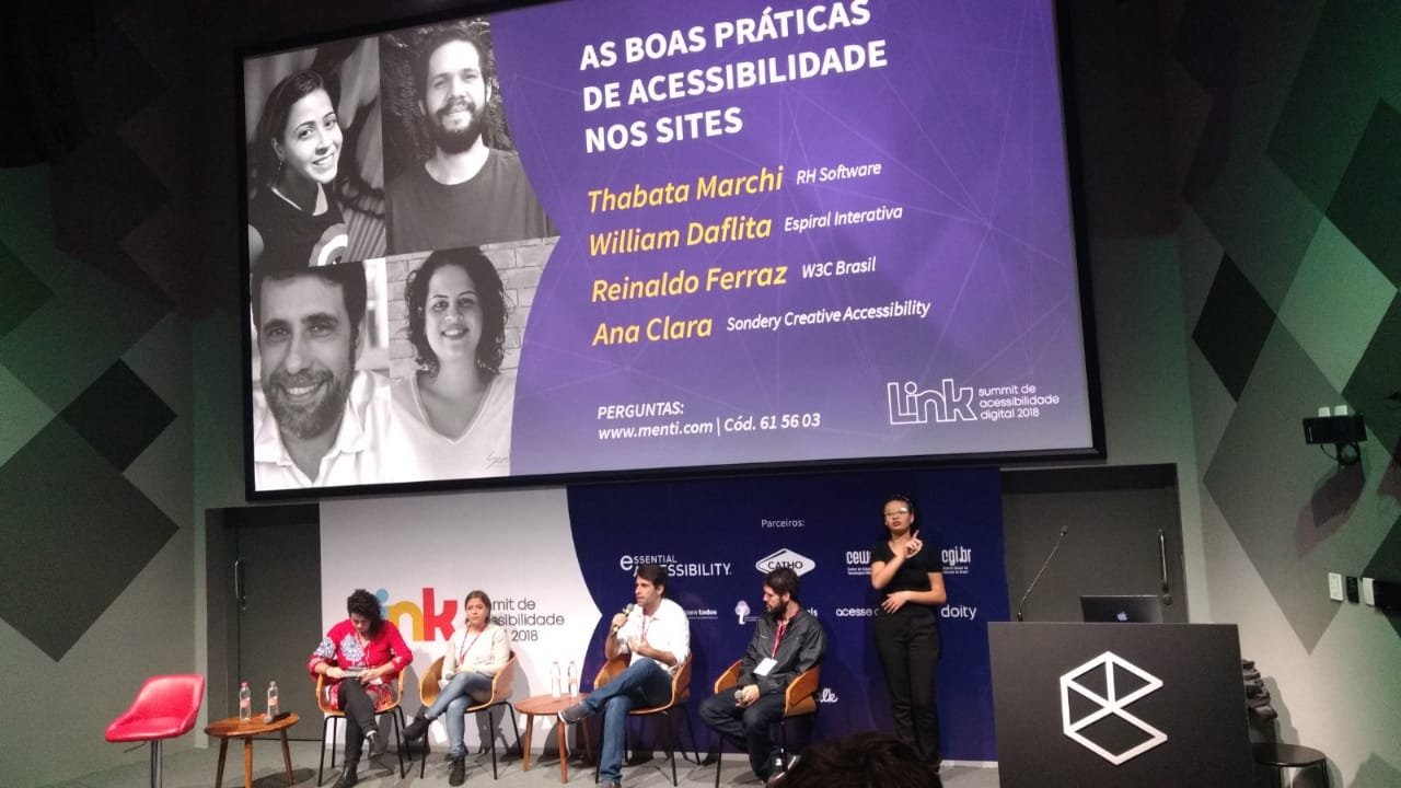 Foto do palco do evento, com um telão com as fotos dos quatro participantes do painel em preto e branco e as seguintes informações sobre um fundo roxo: As boas práticas de acessibilidade nos sites. E os nomes: Thataba Machi, RH Software. William Daflita, Espiral Interativa. Reinaldo Ferraz, W3C Brasil. Ana Clara, Sondery - Creative Accessibility. Sob o telão os quatro participantes estão em cadeiras de madeira, da esquerda para a direita: Ana Clara, uma mulher banca, de cabelos castanhos cacheados com uma lateral raspada usando uma bata vermelha, Thabata Machi, mulher branca de cabelos castanhos lisos amarrados em um rabo de cavalo, usa um casaco branco e uma calça jeans, Reinaldo Ferraz, homem branco de cabelos castanhos, usa camisa branca, calça jeans e segura o microfone na altura da boca e William Daflita, homem branco de cabelo e barba castanho, camisa cinza e calça jeans escura. Ao lado deles, de preto, a intérprete de Libras.