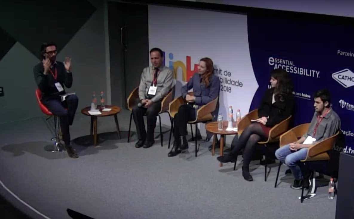 Foto das cinco cadeiras do palco do evento ocupadas pelos palestrantes. Da esquerda para a direita: Alexandre Ohkawa, um homem japonês, de cabelos pretos e roupa preta, Sidney Tobias, um homem moreno com roupa cinza, Fabíola Calixto, uma mulher branca, de cabelos laranja, com um casaco cinza, Bárbara Simão, uma mulher branca de cabelos castanhos cacheados e roupa preta e Gustavo Torniero, um rapaz moreno de cabelos pretos, camiseta cinza e calça jeans.