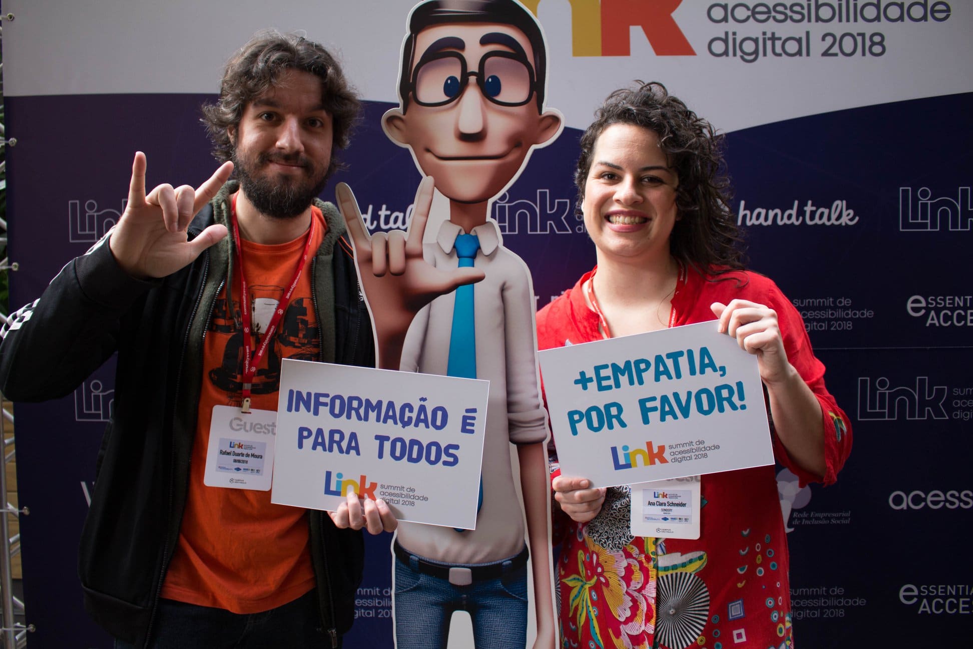 A foto de duas pessoas ao lado do personagem virtual Hugo, em frente a um fundo roxo do evento Link. Na foto estão, da esquerda para a direita: Rafael Duarte, um homem branco de cabelos e barba castanhos, usando um casaco preto e camiseta laranja; em uma das mãos ele segura uma placa escrito “Informação é para todos” e com a outra faz o sinal de “amor” em Libras. Hugo, uma versão impressa do personagem virtual, um homem branco com óculos preto, camisa branca, gravata azul e calça jeans que faz o sinal de “amor” com a mão. Ana Clara, uma mulher branca, de cabelos cacheados raspados na lateral, usa um vestido vermelho estampado e está sorrindo e segurando uma placa que diz “mais empatia, por favor”.