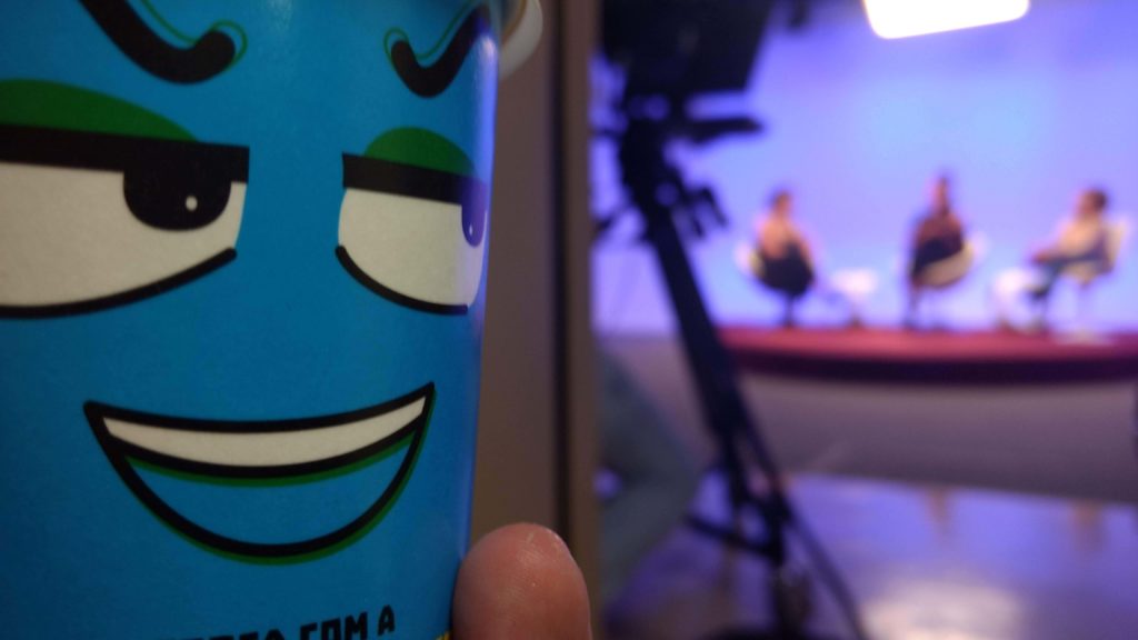 Tomando toda a metade esquerda da foto está um copo de papel azul com um rosto sorrindo desenhado, com olhos virados para a direita. Na metade da direita, que está desfocada, três pessoas estão sentadas em cadeiras brancas sobre um tablado vermelho e em frente a elas, à esquerda, uma câmera filmadora sobre um tripé.