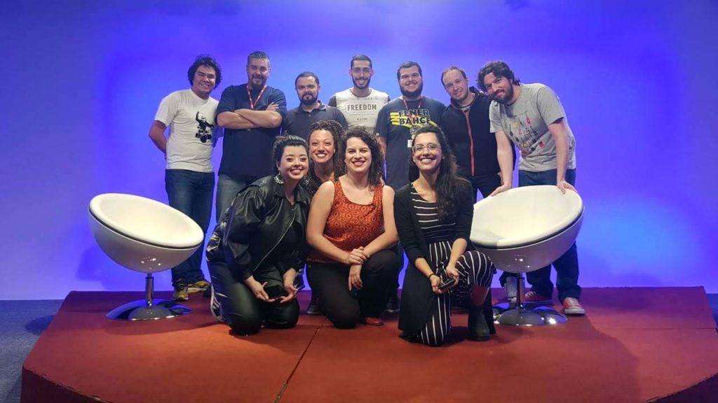 Equipe responsável pela gravação do Mudando de Assunto está de frente, um ao lado do outro, em frente ao cenário do programa: um tablado vermelho com um fundo azul. Todos estão sorrindo. São sete homens em pé atrás e mais adiantadas estão Ana Clara, Erika e mais duas mulheres, que estão entre duas cadeiras brancas com pés metálicos.