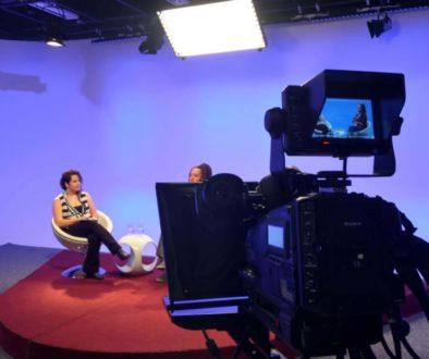 Ana Clara e Erika estão sentadas nas cadeiras brancas do programa, sobre um tablado vermelho e fundo azul. Acima delas estão as luzes de iluminação do estúdio e em frente a elas, à direita da imagem, está uma uma câmera filmadora cujo visor mostra a as duas enquadradas do joelho para cima. Ana Clara usa uma camisa listrada preta e branca e uma calça preta e a Erika está com uma blusa preta e saia marrom.