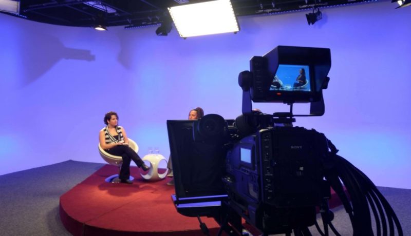 Ana Clara e Erika estão sentadas nas cadeiras brancas do programa, sobre um tablado vermelho e fundo azul. Acima delas estão as luzes de iluminação do estúdio e em frente a elas, à direita da imagem, está uma uma câmera filmadora cujo visor mostra a as duas enquadradas do joelho para cima. Ana Clara usa uma camisa listrada preta e branca e uma calça preta e a Erika está com uma blusa preta e saia marrom.
