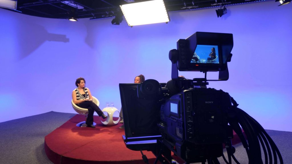 Ana Clara e Erika estão sentadas nas cadeiras brancas do programa, sobre um tablado vermelho e fundo azul. Acima delas estão as luzes de iluminação do estúdio e em frente a elas, à direita da imagem, está uma uma câmera filmadora cujo visor mostra a as duas enquadradas do joelho para cima. Ana Clara usa uma camisa listrada preta e branca e uma calça preta e a Erika está com uma blusa preta e saia marrom.