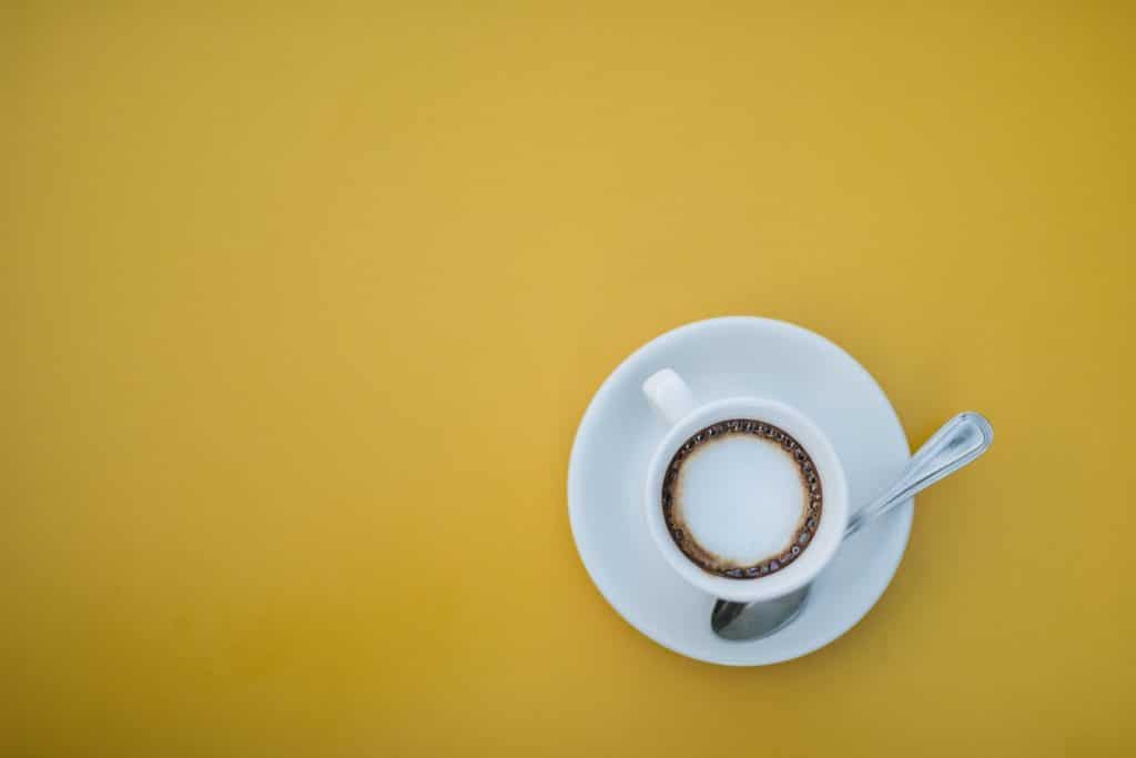 Foto de uma xícara branca de café vista de cima que está sobre um fundo amarelo. A xícara está posicionada na metade inferior direita da imagem, está cheia de café com creme e está apoiada em um pires branco onde também está uma colher prateada.