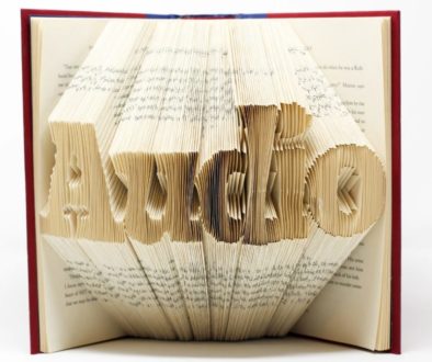 Um livro aberto apoiado em pé em uma superfície branca, as páginas estão dobradas de maneira a formarem a palavra Audio.