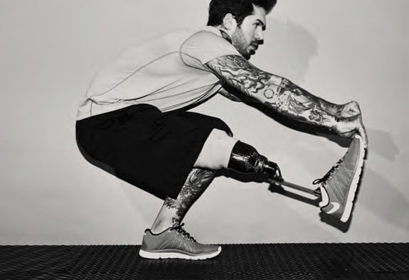 Foto em preto e branco de homem de perfil, virado para a direita com uma prótese do joelho para baixo da perna direita. Ele está agachado, com a perna esquerda flexionada e a direita estendida à frente, tronco inclinado para a frente e segura a ponta do tênis com as mãos. Ele é branco, tem cabelo curto e tatuagens cobrindo ambos os braços e a perna e usa camiseta clara, bermuda preta e tênis esportivo.