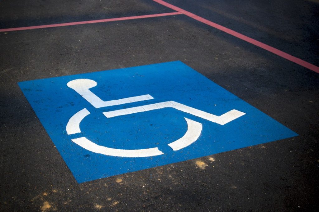 Chão de asfalto com faixas vermelhas pintadas demarcando uma vaga para carros. No meio da vaga está pintado um quadrado azul com um boneco cadeirante branco no centro, o símbolo internacional de acesso.