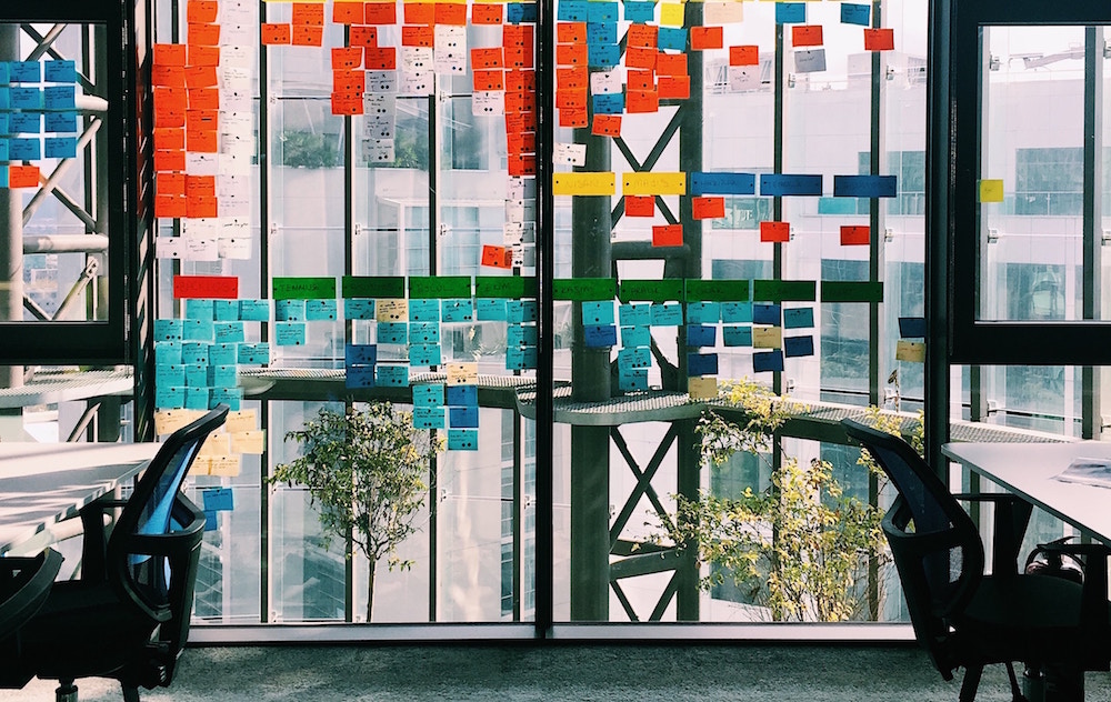 Vista por dentro há uma grande janela de vidro na qual há diversos post-its colados vermelhos, verdes, brancos, azuis e amarelos. Tanto à direita como à esquerda da janela há mesas brancas com cadeiras de escritório pretas encostadas, sobre o chão de carpete cinza.