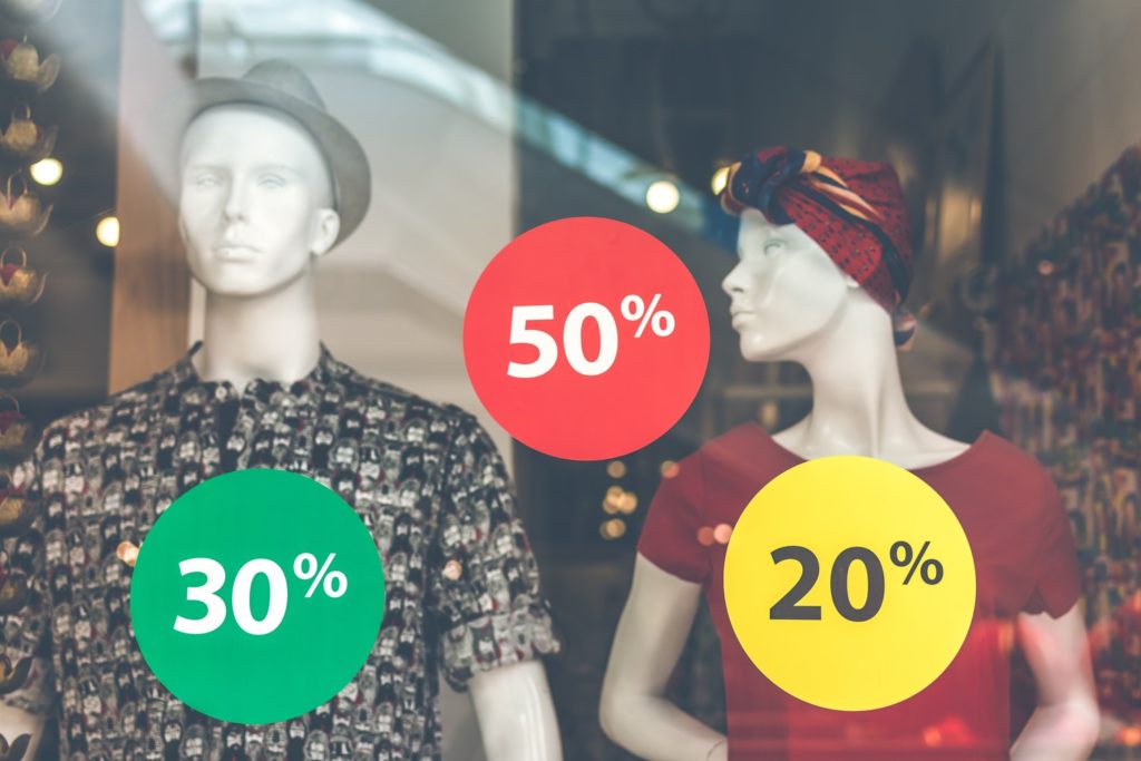 Vitrine de uma loja de roupas. Dois manequins aparecem da cintura para cima. O da esquerda é um manequim masculino, com chapéu cinza e camiseta estampada cinza e preta. À direita um feminino, com lenço vermelho amarrado na cabeça e uma blusa vermelha. Na frente deles, grudados na vitrine, há três adesivos redondos de cores verde, vermelho e amarelo indicando os valores "30%", "50%" e "20%".