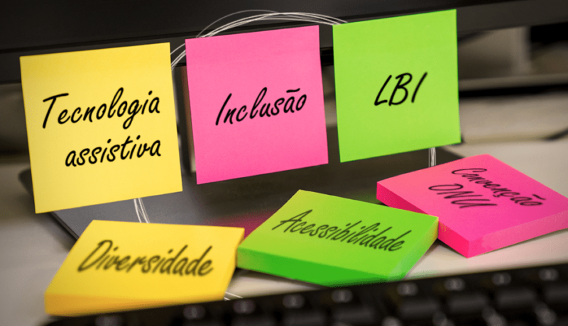 Sobre uma mesa há um computador com três postits nas cores amarelo, rosa e verde grudados. Neles estão escritos em letra cursiva os termos "tecnologia assistiva", "inclusão" e "LBI". Sobre a mesa há três bloquinhos de postits nas mesmas cores, com os termos "diversidade", "acessibilidade" e "convenção ONU".