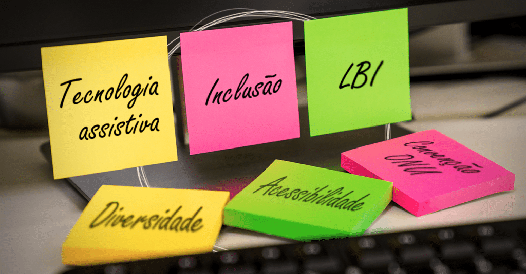 Sobre uma mesa há um computador com três postits nas cores amarelo, rosa e verde grudados. Neles estão escritos em letra cursiva os termos "tecnologia assistiva", "inclusão" e "LBI". Sobre a mesa há três bloquinhos de postits nas mesmas cores, com os termos "diversidade", "acessibilidade" e "convenção ONU".