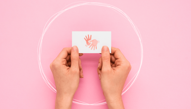 Sobre um fundo rosa claro, duas mãos seguram um cartão branco com um símbolo rosa de duas mãos abertas, uma com os dedos para cima e outra para baixo. Ao redor da imagem há um círculo branco um tracejado como se feito à lápis e reforçado por ele várias vezes.