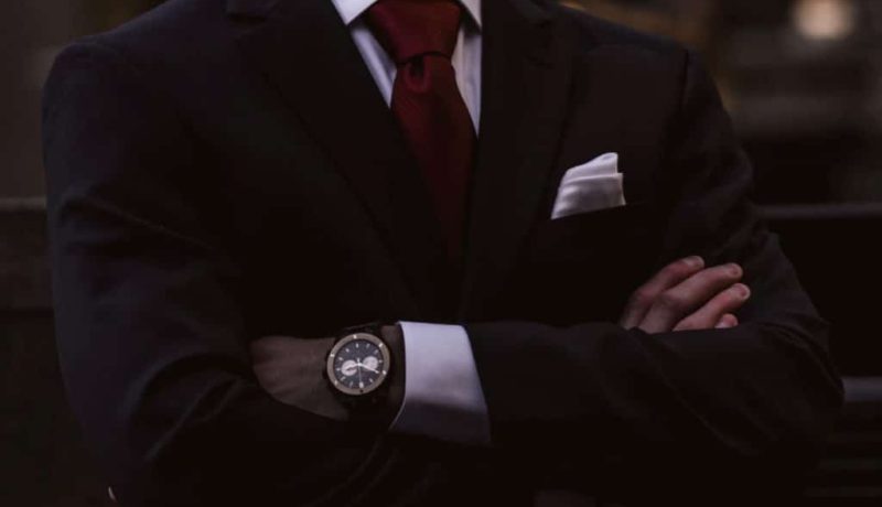 Foto do dorso de um homem de braços cruzados, ele usa um terno preto, grava vermelha e relógio no pulso.