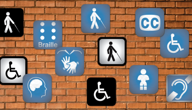 Uma parede de tijolinhos aparentes com várias placas azuis com os símbolos de acessibilidade, como o símbolo internacional do acesso, o símbolo de Braille, o símbolo de pessoa com deficiência visual, símbolo de legendas, símbolo de pessoa com deficiência auditiva, símbolo de pessoa com nanismo.