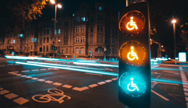 Imagem retangular na horizontal. A imagem é uma foto de uma rua no período da noite. Em primeiro plano, do lado direito da foto, tem um semáforo retangular e vertical preto com três círculos luminosos um embaixo do outro também na vertical. As três luzes - vermelho, amarelo e verde - estão acesas e tem o símbolo de pessoa com deficiência no centro. Em segundo plano é possível ver uma rua com faixas e uma bicicleta na cor branca no asfalto cinza escuro, postes de luz no lado esquerdo, no centro e no lado direito da foto. , uma de cada lado. Também é possível ver pedaços do céu preto nas partes superiores direita e esquerda. Na parte lateral esquerda uma construção bege com várias janelas.