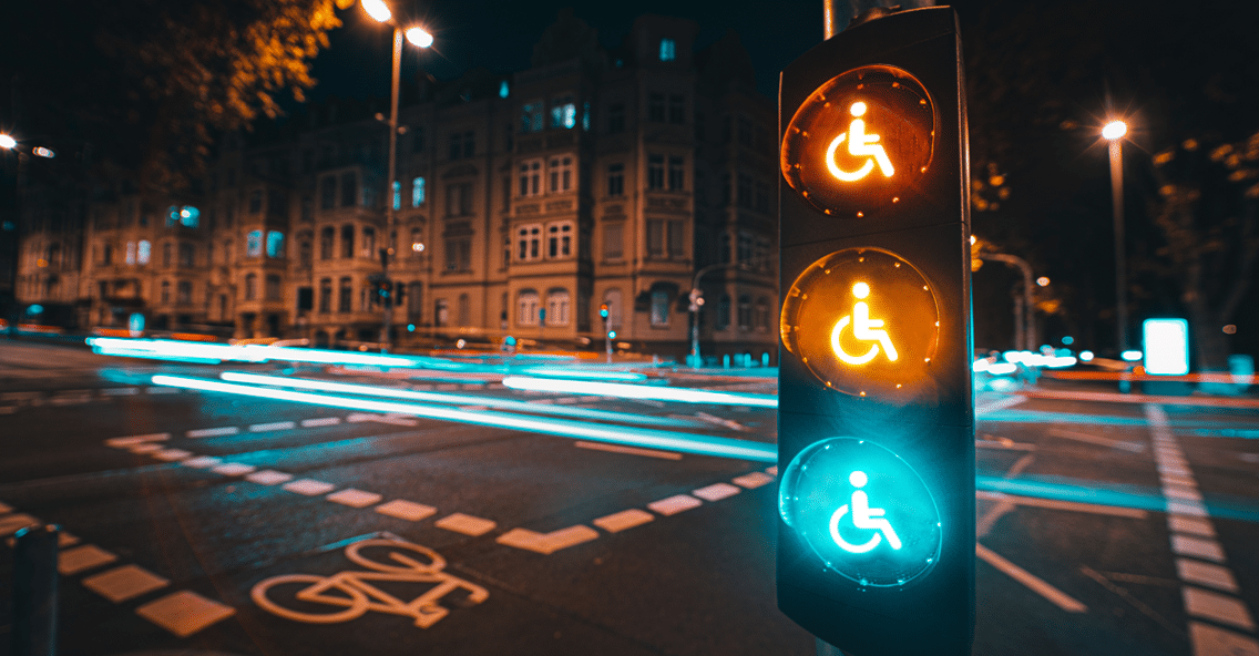 Imagem retangular na horizontal. A imagem é uma foto de uma rua no período da noite. Em primeiro plano, do lado direito da foto, tem um semáforo retangular e vertical preto com três círculos luminosos um embaixo do outro também na vertical. As três luzes - vermelho, amarelo e verde - estão acesas e tem o símbolo de pessoa com deficiência no centro. Em segundo plano é possível ver uma rua com faixas e uma bicicleta na cor branca no asfalto cinza escuro, postes de luz no lado esquerdo, no centro e no lado direito da foto. , uma de cada lado. Também é possível ver pedaços do céu preto nas partes superiores direita e esquerda. Na parte lateral esquerda uma construção bege com várias janelas.