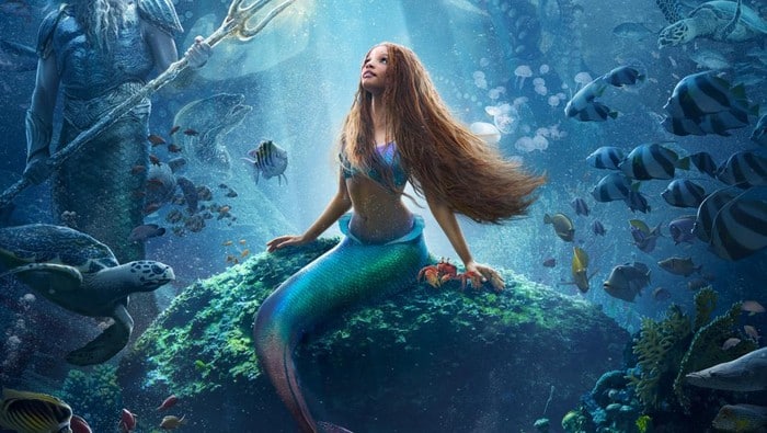 Cena do filme A pequena sereia. Sentada sobre uma pedra no fundo do mar, cercada por peixes, há uma sereia. Ela é negra, tem longos cabelos marrom claro, usa um top roxo e tem uma cauda verde. Ao lado dela há um caranguejo.